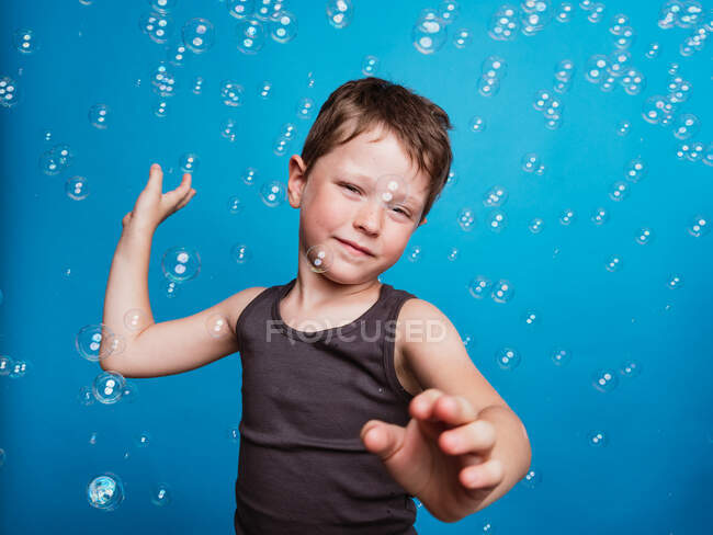 Niño preadolescente mirando a la cámara en el estudio con burbujas de jabón volador sobre fondo azul - foto de stock