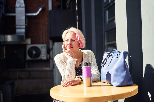 Heitere ungezwungene Frau sitzt am Tisch mit umweltfreundlicher Tasse mit Heißgetränk in der Stadt an einem sonnigen Tag — Stockfoto