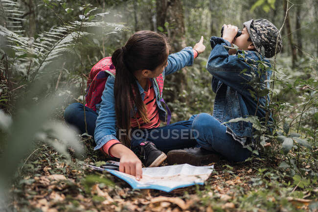 Ethnic girl showing green plant to brother while looking away on binocular and sitting on terrain with paper map and loupe in woods — Stock Photo