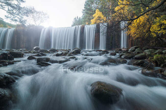 Vista panorámica de la cascada que desciende por rocas en bosques montañosos en otoño en larga exposición en el río Lozoya en el Parque Nacional Guadarrama - foto de stock