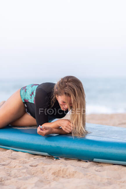 Vista lateral del surfista femenino positivo en traje de baño acostado en el paddleboard en la orilla arenosa contra el mar y mirando hacia otro lado - foto de stock