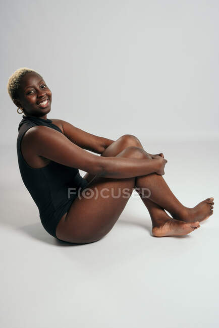 Heureuse femme afro-américaine en body noir et avec corps courbé assis avec jambes croisées en studio sur fond gris et regardant la caméra — Photo de stock