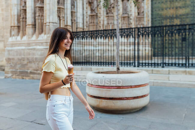 Cultiver joyeuse jeune femme en pendentif et boucles d'oreilles avec de délicieux gelato en cône de gaufre regardant loin dans la rue — Photo de stock
