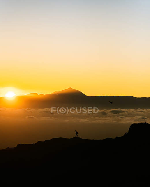 Удаленный обзор силуэта туриста, идущего вдоль скалистой горной цепи против закатного неба с оранжевым солнцем — стоковое фото