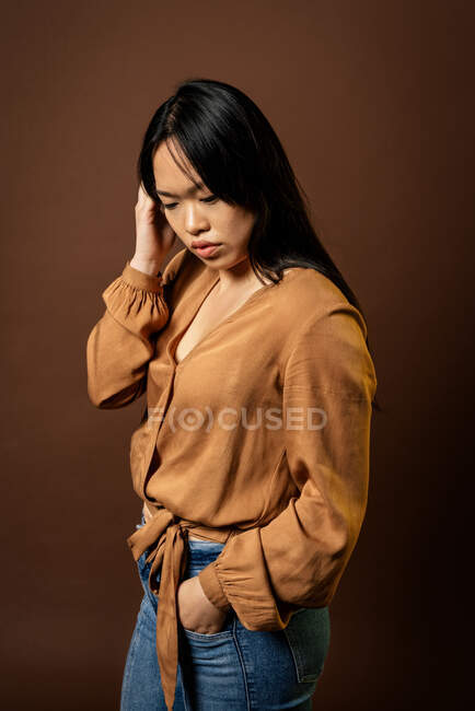 Слайд-вид азиатской женщины в трикотажной одежде, смотрящей вниз на коричневом фоне в студии — стоковое фото