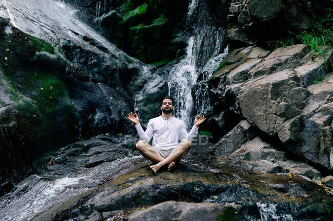 Peaceful male sitting in Padmasana with mudra hands and closed eyes while doing yoga and meditating on wet rock near waterfall — Stock Photo