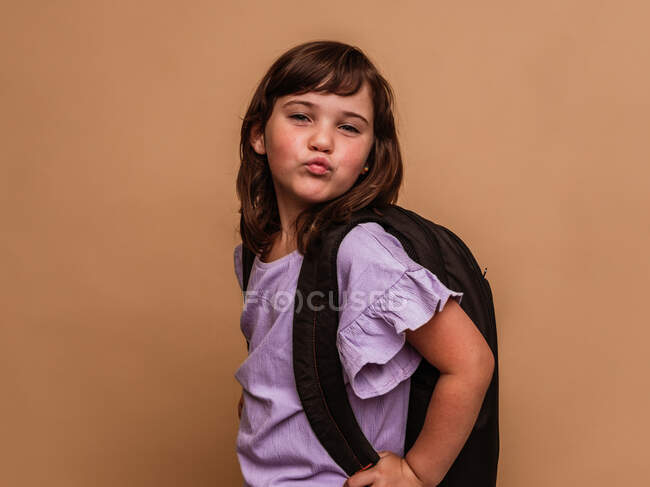 Cute schoolchild with rucksack pouting lips while standing on brown background in studio and looking at camera — Stock Photo