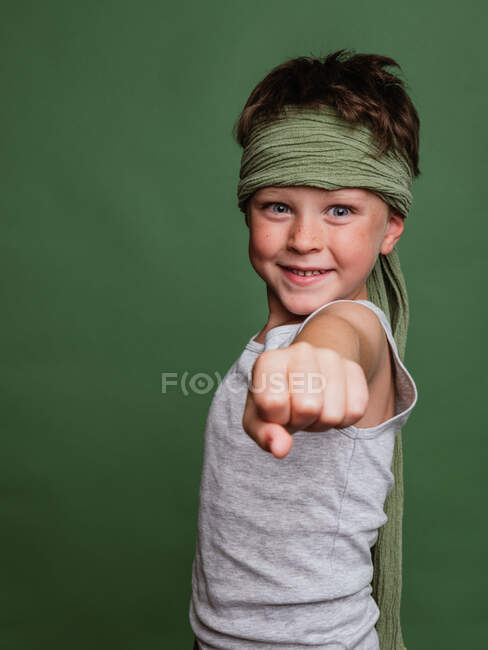Cheerful preteen karate boy in hachimaki headscarf and with outstretched clenched fist looking at camera on green background in studio and having fun — Stock Photo