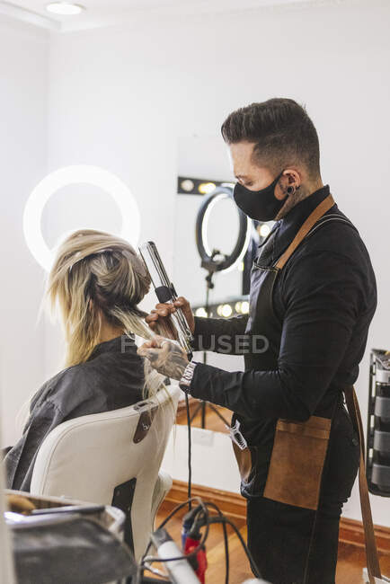 Мужчина-парикмахер, использующий утюг для завивки блондинистых замков женщины-клиента во время работы в салоне красоты — стоковое фото