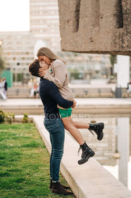 Вид сбоку любящего бойфренда, поднимающего стильную подругу во время поцелуя на городской улице — стоковое фото