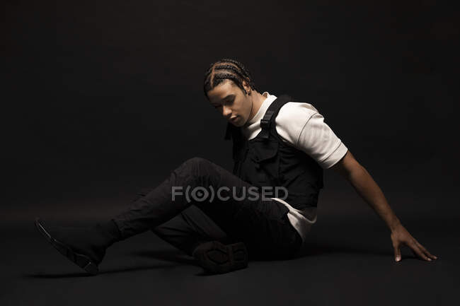 Side view of confident young ethnic hipster male model with braided hair wearing white shirt with black clothes looking down while sitting in dark studio — Stock Photo