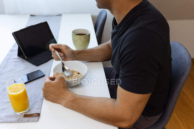 Vista lateral da cultura macho anônimo comer granola, tendo café da manhã saudável e sentado à mesa com um copo de suco de laranja e gadgets em casa — Fotografia de Stock