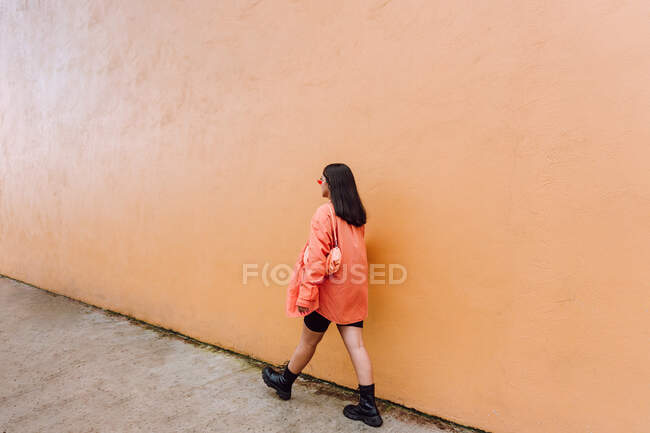 Ganzkörperrückansicht einer stilvollen Frau in trendigem Outfit und Sonnenbrille, die an orangefarbener Wand vorbeiläuft — Stockfoto