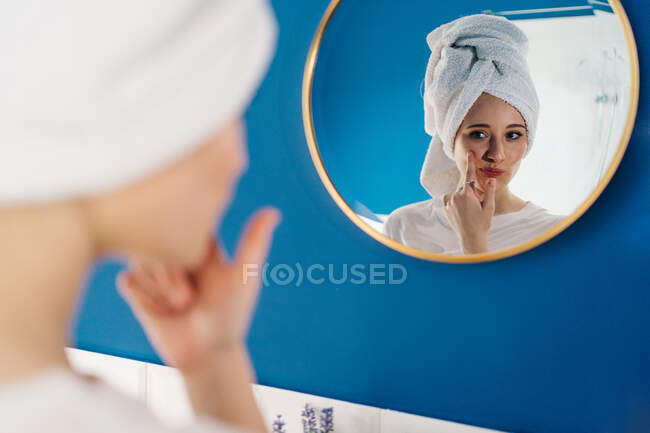 Visão traseira da fêmea em turbante de toalha refletindo no espelho no banheiro e aplicando creme facial durante a rotina de cuidados com a pele de manhã — Fotografia de Stock