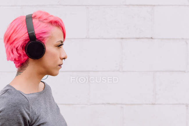 Side view of young female with bright pink hair listening to music with headphones while standing near white wall — Stock Photo