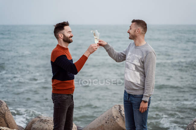 Homosexual male partners with modern haircuts enjoying champagne from glasses while standing on ocean coast in daytime — Stock Photo