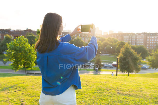 Вид ззаду на стильну жінку, що стоїть на зеленому трав'яному пагорбі і фотографує міський пейзаж на смартфоні в сонячний день — стокове фото