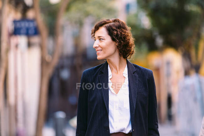 Позитивна жінка в стильному одязі, що йде по вулиці посміхаючись дивиться вбік — стокове фото