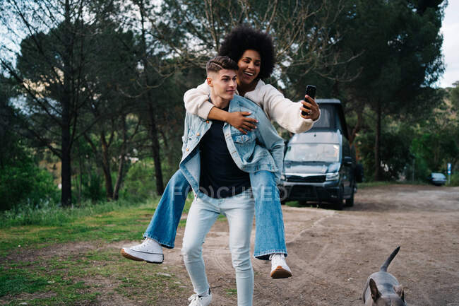 Улыбающаяся чёрная женщина, катающаяся на спине с парнем во время автопортрета на мобильном телефоне против собаки и каравана в лагере — стоковое фото