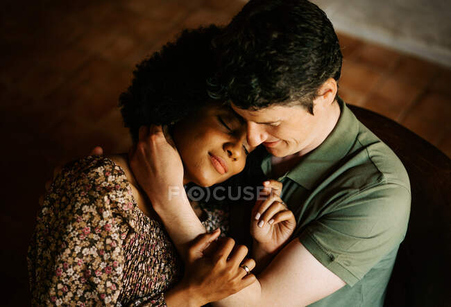 Сверху любящая черная женщина с закрытыми глазами, сидящая на коленях у мужчины, обнимаясь дома — стоковое фото