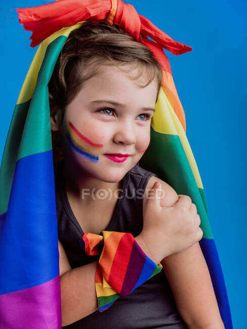 Niña con bandera de arco iris atada en la cabeza mirando hacia otro lado mientras está de pie sobre fondo azul - foto de stock