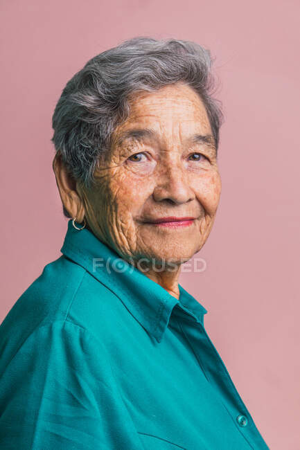 Vue latérale de la femelle âgée aux cheveux gris courts et aux yeux bruns regardant la caméra sur fond rose en studio — Photo de stock