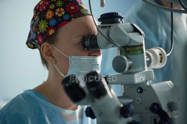 Médecin adulte ciblé en masque stérile et coiffe médicale ornementale regardant au microscope chirurgical contre un collègue de culture à l'hôpital — Photo de stock