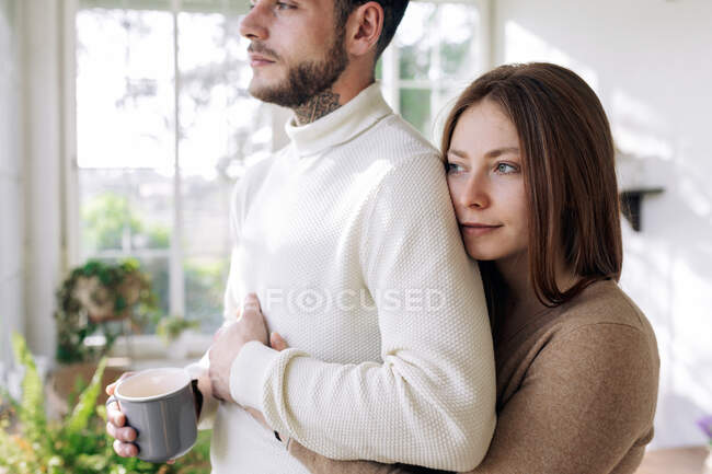 Vista lateral de la alegre hembra abrazando al macho barbudo amado con taza de café mientras mira hacia otro lado contra la ventana en casa - foto de stock