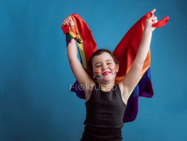 Ragazza sorridente con guancia dipinta alzare le braccia con bandiera multicolore su sfondo blu vivido — Foto stock