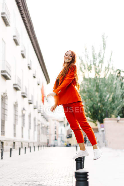 Side view of redhead female in bright orange suit standing on metal parking bollard in street and looking at camera — Stock Photo