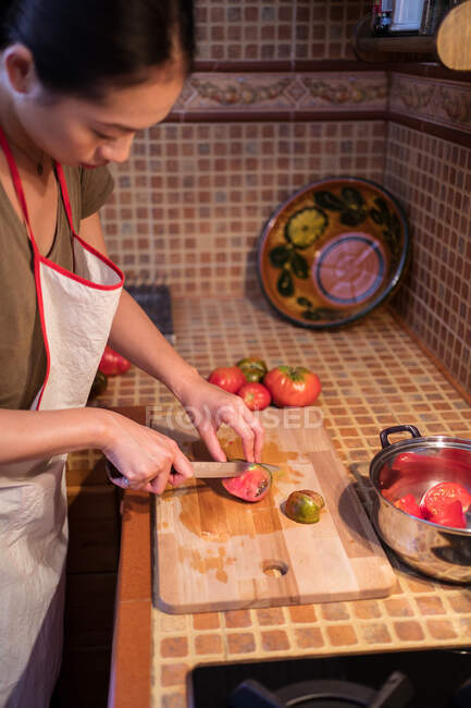 Da sopra vista laterale di femmina etnica in grembiule tagliando pomodori maturi sul tagliere mentre si cucina il pranzo in cucina a casa — Foto stock