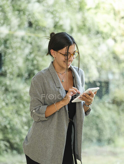 Feminino em óculos usando tablet enquanto está de pé em vidro de local de trabalho espaçoso com verde atrás e trabalhando no projeto — Fotografia de Stock