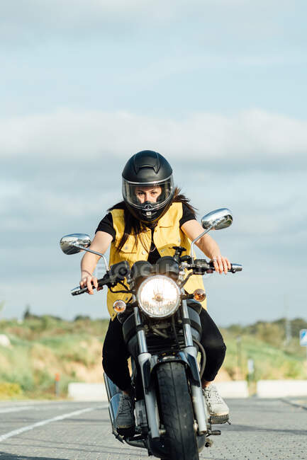 Bicicleta feminina focada no capacete andando de moto moderna ao longo da estrada no dia ensolarado e olhando para a câmera — Fotografia de Stock