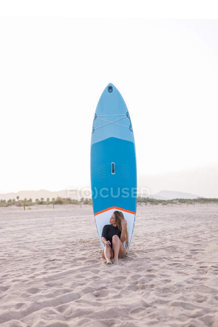 Female surfer sitting with blue SUP board on sandy seashore in summer and looking away — Stock Photo