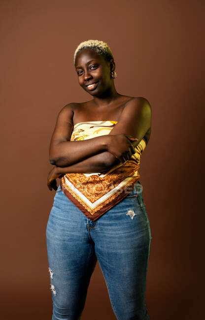 Modelo feminino afro-americano feliz vestindo top cachecol de seda com braços dobrados em fundo marrom em estúdio e olhando para a câmera — Fotografia de Stock