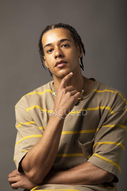 Joven modelo masculino afroamericano reflexivo con cabello trenzado vestido con una camisa de rayas de gran tamaño y un collar mirando a la cámara contra el fondo gris - foto de stock