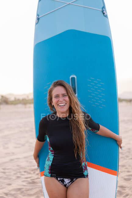 Glückliche Surferin steht im Sommer mit blauem SUP-Board am Sandstrand und blickt in die Kamera — Stockfoto