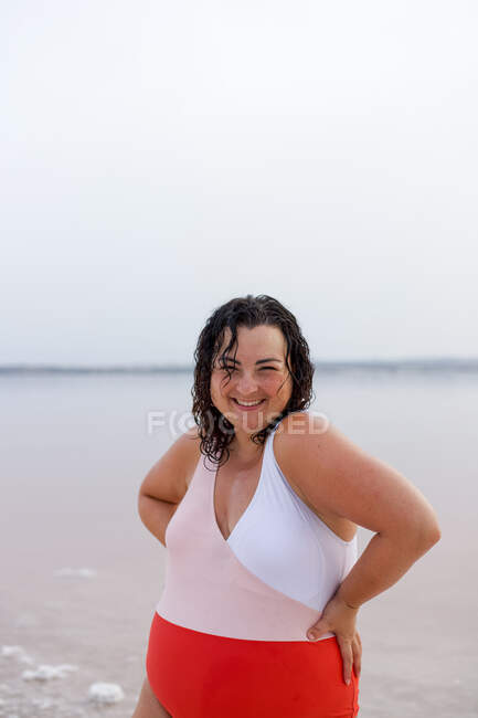 Positive kurvige Frau im Badeanzug steht mit den Händen auf der Taille am Strand in der Nähe des rosa Teiches und schaut in die Kamera, während sie den Sommerurlaub genießt — Stockfoto