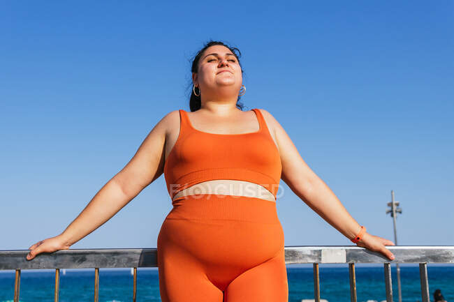 Concentrated ethnic female athlete with curvy body and closed eyes in active wear against fence and ocean in sunlight — Stock Photo