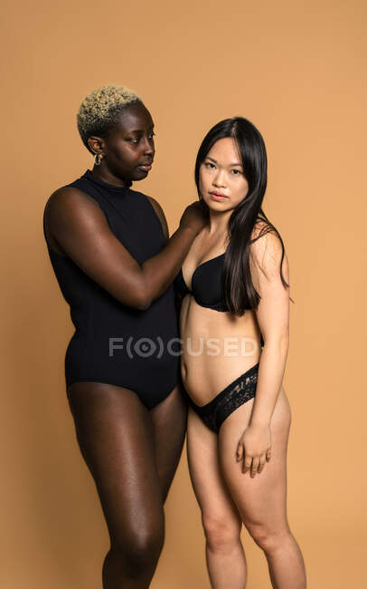 Modelos femeninos multiraciales en lencería negra con una mano en el hombro de la otra sobre fondo beige para concepto de positividad corporal en estudio - foto de stock