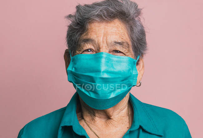 Sorridente femmina anziana utilizzando blu maschera medica protettiva da coronavirus mentre guardando la fotocamera su sfondo rosa in studio — Foto stock
