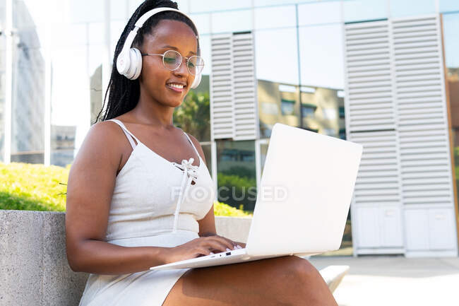Mujer afroamericana positiva en auriculares sentada con portátil en la calle en Barcelona y disfrutando de canciones mientras mira la pantalla - foto de stock