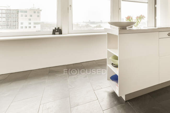 Innendetails der modernen geräumigen Loft-Wohnung mit weißer Küchentheke mit dekorativen Tellern vor dem Panoramafenster mit Fernglas auf der Fensterbank — Stockfoto