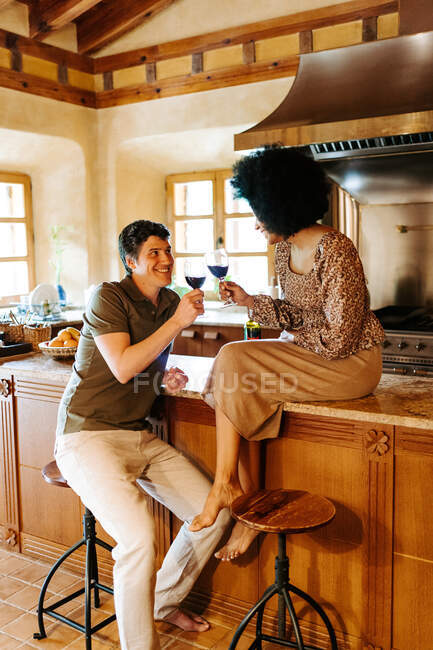 Delighted black woman sitting on counter and man sitting on stool in kitchen clinking glasses with alcohol drink while celebrating event at home and looking at each other — Stock Photo