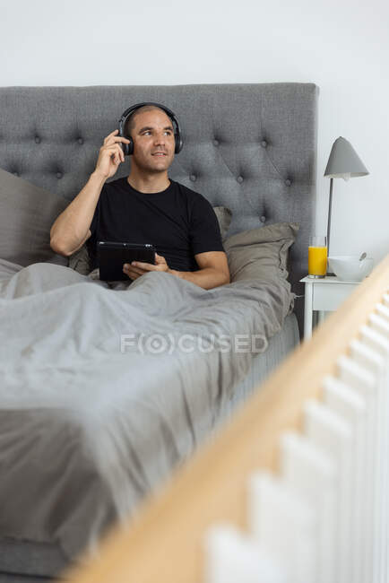 Gelassener Mann mit Kopfhörer sitzt auf dem Bett unter einer Decke und surft auf dem Tablet in den sozialen Medien, während er morgens Musik hört — Stockfoto