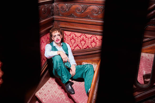 From above of eccentric male actor with smeared makeup sitting on staircase during performance — Stock Photo