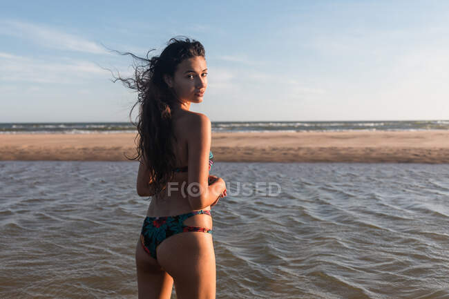 Entzückte Frau im Badeanzug steht am nassen Strand am Meer und blickt in die Kamera, während sie den Sommerurlaub genießt — Stockfoto