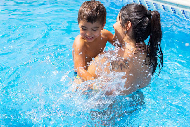 Back view of unrecognizable mother with cheerful son standing in swimming pool near green trees on sunny summer day — Stock Photo