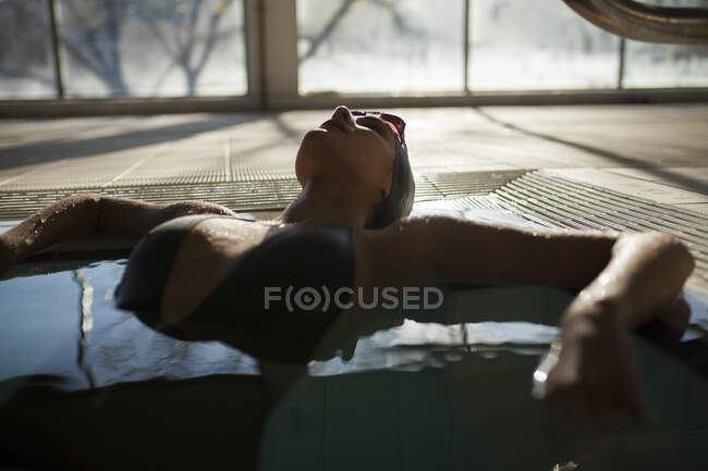 Jeune belle femme allongée sur le bord de la piscine intérieure, portant un maillot de bain noir — Photo de stock