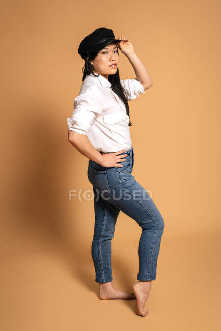 Сторона огляду моди азіатська жіноча модель в білій сорочці і джинсах торкається шапки, стоячи на бежевому фоні і дивлячись на камеру — стокове фото